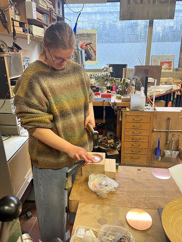 Person mit Brille und Pferdeschwanz hämmert konzentriert auf ein Metallwerkstück, das auf einem Holzblock liegt. Die Umgebung ist eine helle, aufgeräumte Werkstatt mit Werkbänken und Regalen.