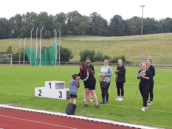 Unser Motto zum Sportfest: "Auf zu neuen Galaxien!" Eine Siegerehrung auf einem Sportplatz: Ein Kind bekommt eine Medaille überreicht, während mehrere junge Frauen mit Medaillen und einem Mikrofon bereitstehen.
