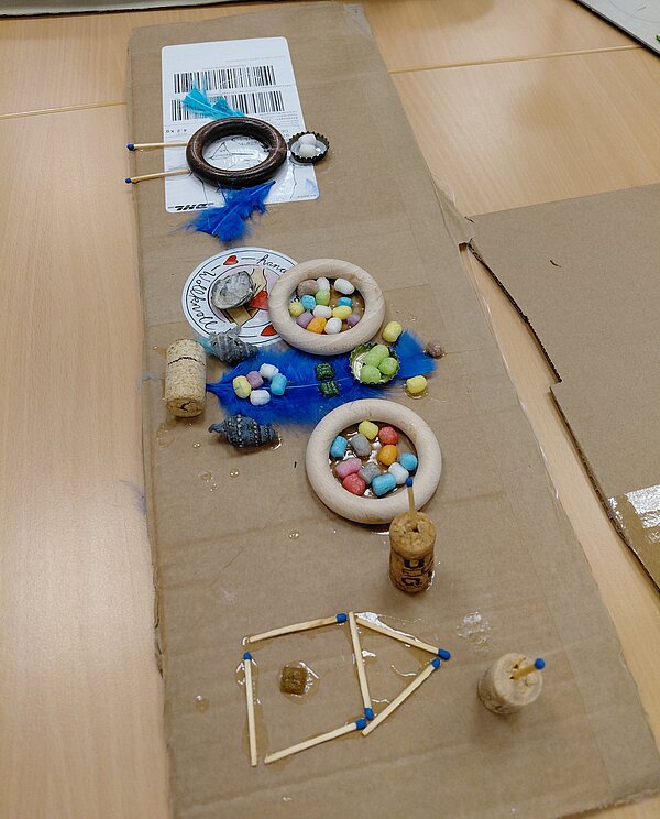 Eine längliche Kartonplatte ist als Collage gestaltet: Holzringe mit bunten Kügelchen, blaue Federn, Korken, Muscheln und ein aus Streichhölzern gelegtes Häuschen sind darauf mit Kleber befestigt.