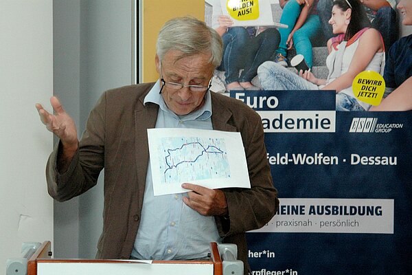 Ein älterer Mann mit Brille hält am Rednerpult eine Ansprache und zeigt dabei eine Zeichnung. Hinter ihm steht ein Roll-up der Euro Akademie Bitterfeld-Wolfen und Dessau.