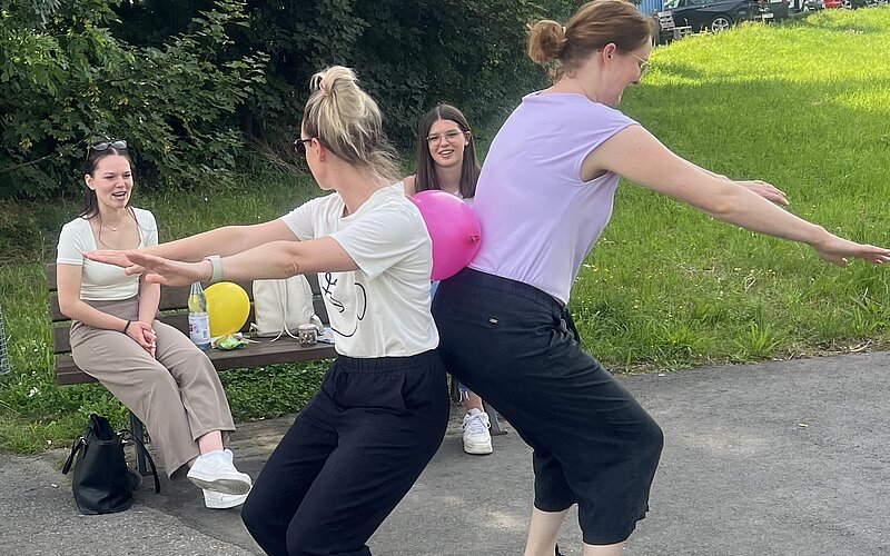 Eine Gruppe von Menschen im Freien, die an einer Aktivität mit Ballons teilnehmen. Zwei Personen im Vordergrund halten einen Ballon zwischen sich, während andere auf einer Bank sitzen und zuschauen.