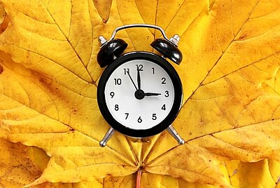 Ein schwarzer Wecker, der auf drei Uhr steht, liegt auf herbstlich gelbem Ahornblatt.