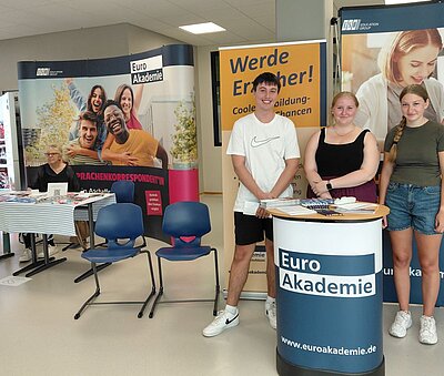 Drei junge Personen stehen lächelnd hinter einem Infotisch der Euro Akademie. Im Hintergrund sind Roll-up-Banner mit Werbung für die Euro Akademie und dem Slogan „Werde Erzieher! Coole Ausbildung – Top Chancen“ zu sehen. Auf dem Tisch liegen Flyer und Informationsmaterialien aus. Links im Bild sitzt eine Person an einem Tisch mit weiteren Materialien.