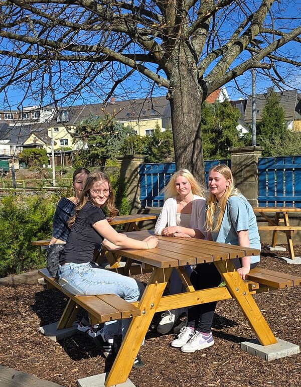 Vier Schülerinnen sitzen draußen im Grünen Klassenzimmer der Euro Akademie Hohenstein-Ernstthal an Holztisch und auf der Holzbank und posieren gut gelaunt für ein Foto.