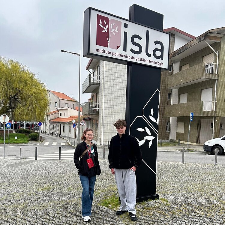 Zwei Auszubildende der Euro Akademie Berlin stehen vor einem Schild mit der Aufschrift „isla instituto politécnico de gestão e tecnologia“. Das hohe Schild zeigt das Logo und den Namen des Instituts in portugiesischer Sprache. Die Szenerie zeigt ein Stadtgebiet mit Gebäuden und Straßenschildern im Hintergrund. Die Azubis sind leger gekleidet: Die Person links trägt Jeans und eine Jacke, die Person rechts eine Jogginghose und eine Jacke.
