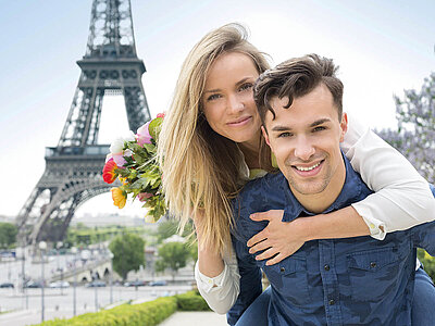 Zwei Personen stehen vor dem Eiffelturm in Paris. Die eine Person hält einen farbenfrohen Blumenstrauß, während die andere sie liebevoll umarmt. Der Himmel ist klar, und die Szenerie strahlt Romantik aus.