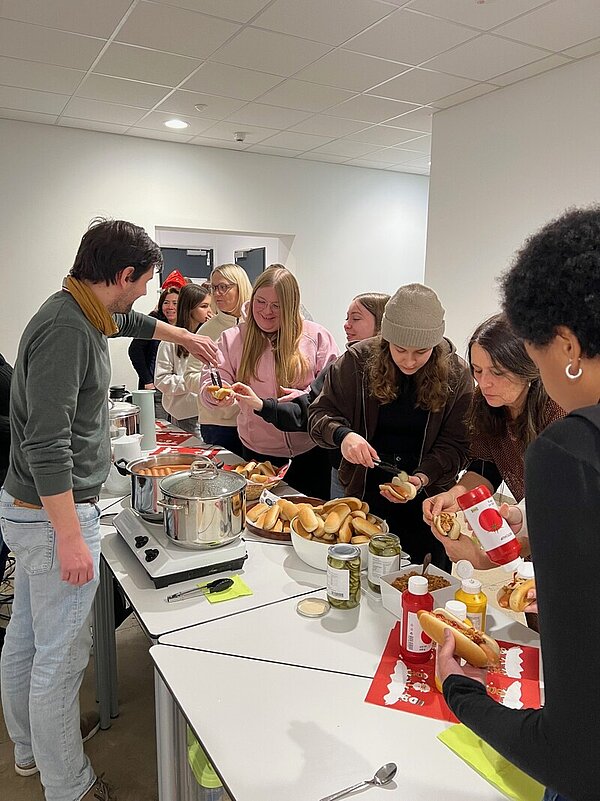 Im Flur drängen sich die Schülerinnen und Schüler entlang der Tische, auf denen die Hot Dog-Zutaten angerichtet sind: Töpfe mit heißen Würstchen, weichen Brötchen, Gurkenscheiben in Gläsern, Röstzwiebel, Ketchup und Senf. Ein Schüler legt einer Schülerin gerade ein Wurst in ihr Brötchen.