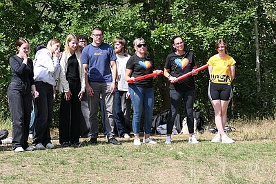 Die Gruppe der Schülerinnen und Schüler steht auf einer Wiese vor einem Hintergrund aus grünen Bäumen. Zwei Personen in der Mitte halten jeweils einen roten Gegenstand in den Händen. Die Kleidung ist überwiegend sportlich, darunter T-Shirts, Hosen und Shorts. Die Stimmung wirkt entspannt, einige lächeln. Im Hintergrund liegen Taschen und Kleidungsstücke auf dem Boden.