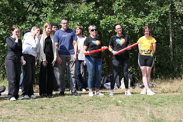Die Gruppe der Schülerinnen und Schüler steht auf einer Wiese vor einem Hintergrund aus grünen Bäumen. Zwei Personen in der Mitte halten jeweils einen roten Gegenstand in den Händen. Die Kleidung ist überwiegend sportlich, darunter T-Shirts, Hosen und Shorts. Die Stimmung wirkt entspannt, einige lächeln. Im Hintergrund liegen Taschen und Kleidungsstücke auf dem Boden.
