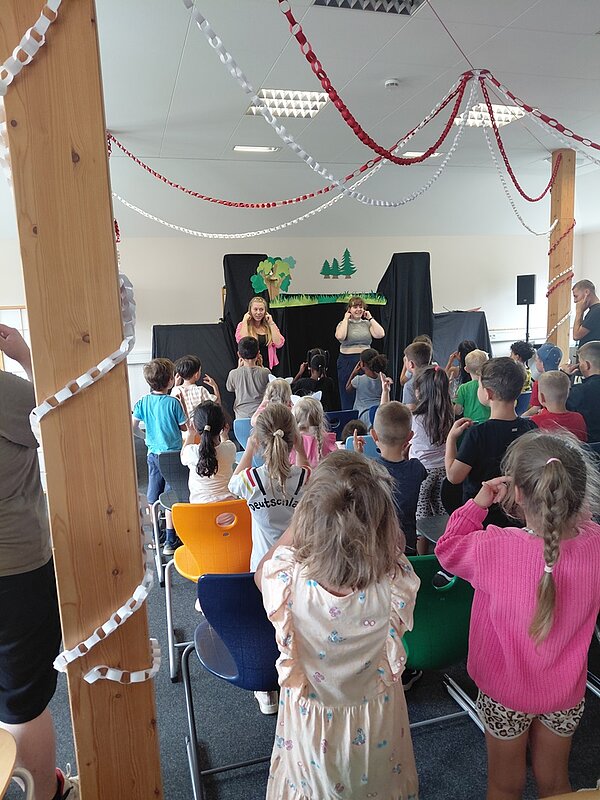 Kinder stehen in einem Raum mit bunten Stühlen und blicken auf eine kleine Bühne, auf der zwei Erwachsene ein Puppentheater aufführen. Die Decke ist mit roten und weißen Papiergirlanden geschmückt. Im Hintergrund sind ein schwarzer Vorhang und grüne Dekorationen in Form von Bäumen zu sehen. Einige Kinder heben die Hände, als würden sie Bewegungen nachmachen oder mitmachen.