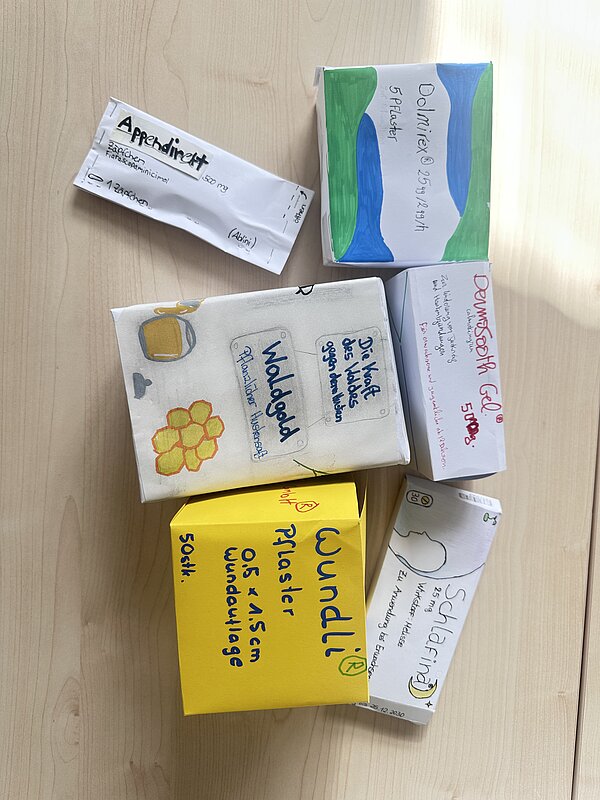 Draufsicht auf mehrere selbst gebastelte Arzneimittelverpackungen auf einem Holztisch. Die Modelle tragen unterschiedliche Produktnamen und Angaben zu Pflastern, Gel, Hustensaft und weiteren Medikamenten.