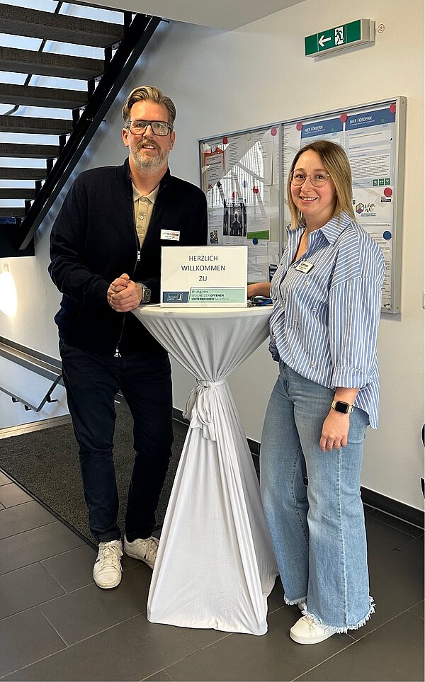 SCHAU REIN! – Woche der offenen Unternehmen Sachsen Ein Mann und eine Frau stehen an einem Stehtisch im Eingangsbereich eines Gebäudes. Auf dem Tisch steht ein Schild mit der Aufschrift „Herzlich willkommen zu“ – vermutlich ein Empfang für eine Veranstaltung.