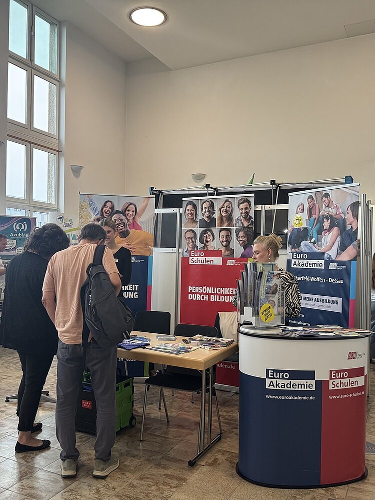 Messestand der Euro Akademie mit zwei nebeneinanderstehenden Theken, die mit den Logos und Farben von Euro Akademie und Euro Schulen gestaltet sind. Auf den Theken liegen Prospekte und Informationsmaterialien. Im Hintergrund hängen große Plakate mit Gruppenfotos und Slogans. Zwei interessierte Personen stehen vor dem Stand und sprechen mit einer Mitarbeiterin, die hinter der Theke steht. Die Szene spielt in einem hellen Raum mit hohen Fenstern und gefliestem Boden.