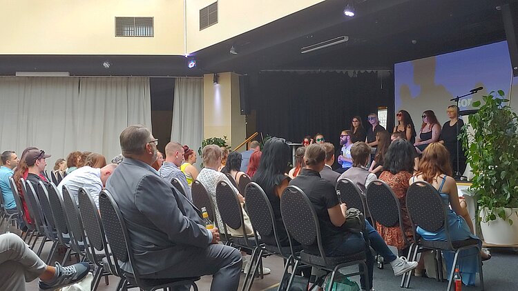 Teilnehmer einer feierlichen Veranstaltung sitzen in Reihen vor einer Bühne. Auf der Bühne stehen mehrere Personen, zum Teil mit Sonnenbrillen, vor einer Leinwand und sprechen oder präsentieren. Im Vordergrund und am Rand stehen große grüne Pflanzen. Die Veranstaltung wirkt wie eine Zeugnisübergabe oder Abschlussfeier.