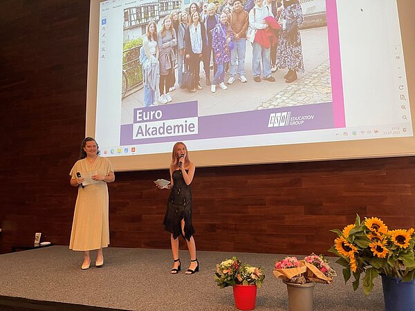 Zwei festlich gekleidete Frauen stehen auf einer Bühne und halten ein Mikrophon in der einen und einen Zettel in der anderen Hand, wobei eine von ihnen ins Mikrophon spricht. Im Hintergrund ist auf einem großen Screen ein Gruppenfoto zu sehen mit dem Logo der Euro Akademie. Auf der Bühne stehen mehrere Blumensträuße in Eimern.