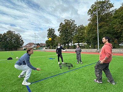 Zwei Personen spielen auf einem Kunstrasenplatz Spikeball. Ein Spieler im Vordergrund beugt sich tief, um den Ball zu spielen, während drei weitere Personen um das Netz herumstehen. Im Hintergrund sind Bäume und ein Sportplatz zu sehen.