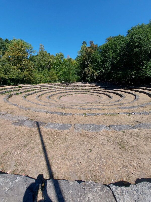 Ein spiralförmiges Muster, das in einen steinernen Boden eingearbeitet ist, umgeben von grünen Bäumen und einem blauen Himmel.