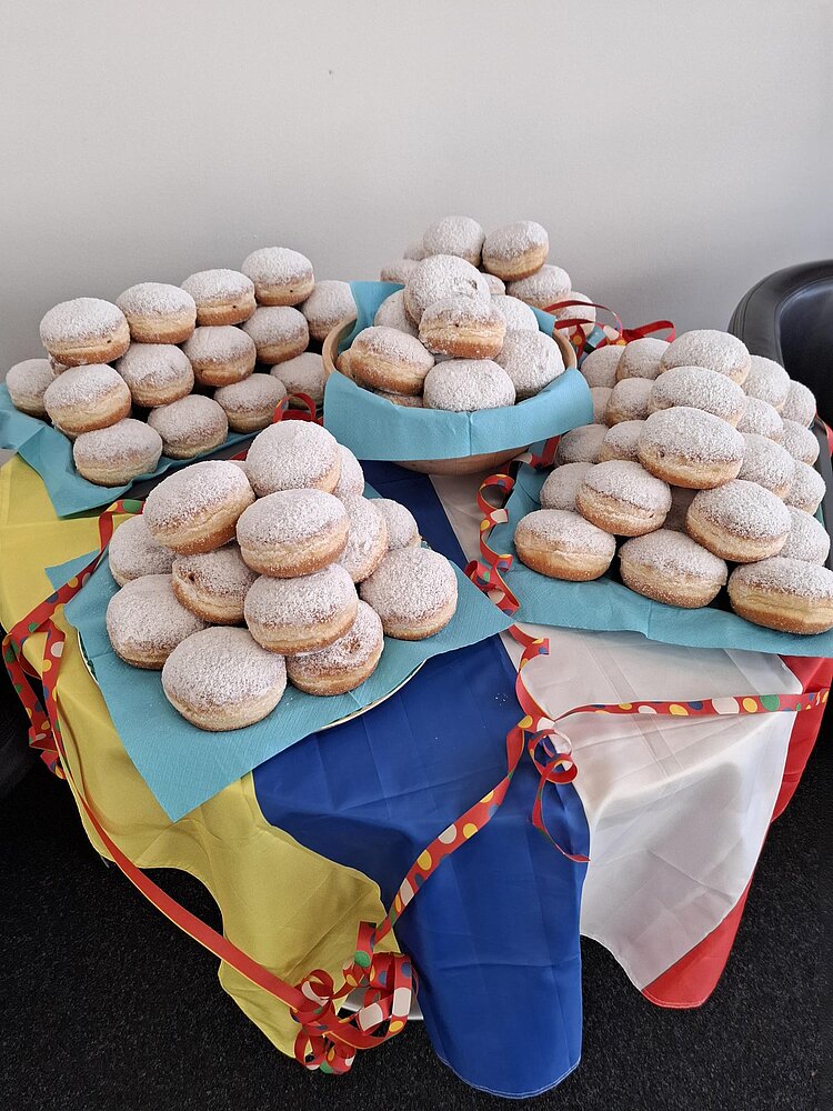 Die Euro Akademie Mainz wünscht tolle Fastnachtstage! Auf einem Tisch mit eine blau, gelb, rot und weißen Fahne liegen viele Berliner mit Puderzucker auf Tellern gestapelt.