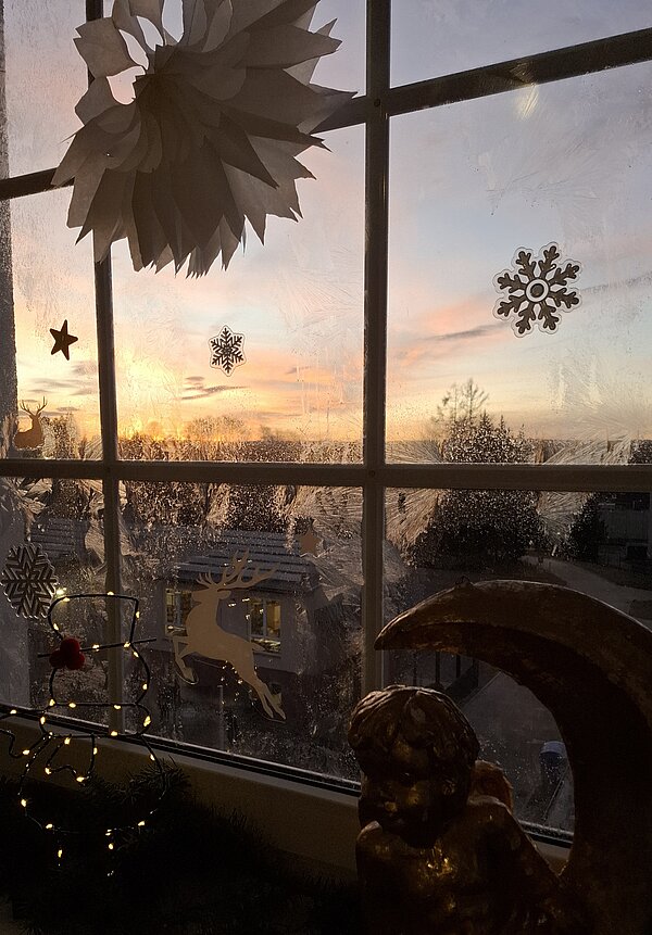 Ein dekoriertes Fenster mit Schneeflocken- und Sternaufklebern, dahinter ein farbenfroher Sonnenuntergang. Am Fensterbrett stehen eine Engelsfigur und weihnachtliche Lichter.