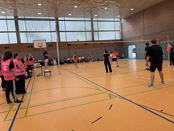 In einer Sporthalle mit gelbem Boden und hohen Fenstern findet ein Volleyballspiel statt. Zwei Mannschaften stehen sich auf dem Spielfeld gegenüber, einige Personen tragen orangefarbene Leibchen, andere schwarze Sportkleidung. Am Spielfeldrand sitzen und stehen weitere Personen, einige tragen pinkfarbene Westen mit weißer Rückennummer. Ein Schiedsrichter steht auf einem Podest neben dem Netz. Im Hintergrund sind Zuschauer auf Bänken zu sehen.