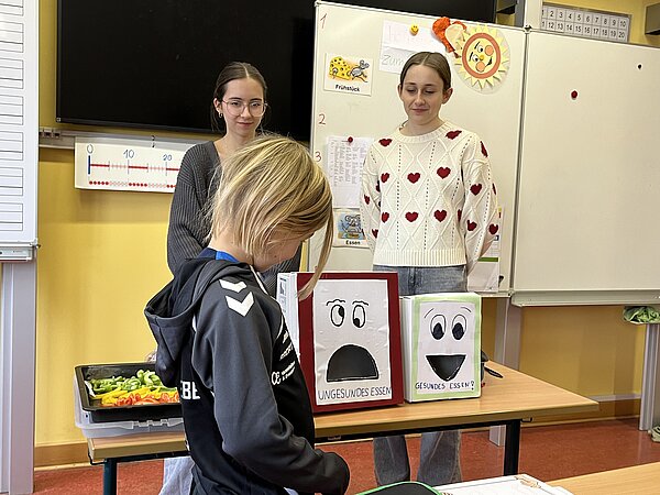 Kind steht an einem Tisch vor zwei selbstgebastelten Boxen mit Gesichtern und den Beschriftungen ‚Ungesundes Essen‘ und ‚Gesundes Essen‘; im Hintergrund stehen zwei Personen und eine Schale mit Gemüsestreifen.