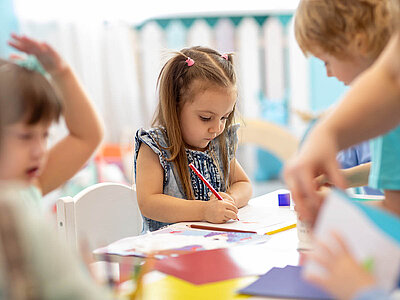Ein Mädchen mit langen Haaren und einer rosa Haarspange sitzt an einem Tisch und malt konzentriert mit einem roten Stift auf einem Blatt Papier. Um sie herum sind weitere Kinder, die ebenfalls beschäftigt sind. Der Raum wirkt hell und kinderfreundlich.