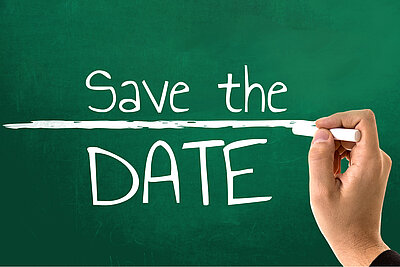Die Aufschrift "Save the Date" ist wie mit weißer Kreide geschrieben auf einer grünen Tafel zu sehen, während eine Hand mit Kreide eine Linie zwischen den Wörtern "Save the" und "Date!" zieht.