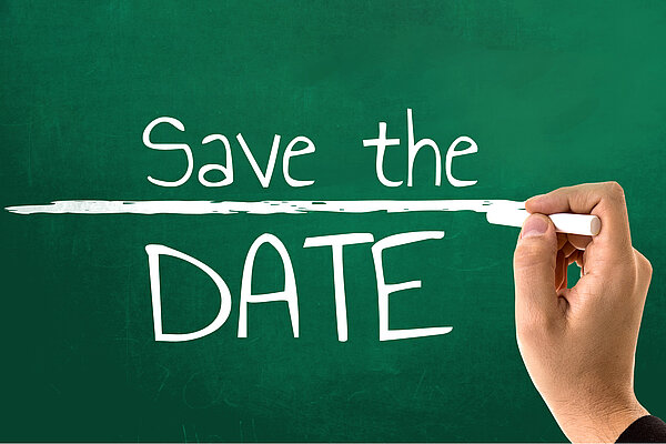 Die Aufschrift "Save the Date" ist wie mit weißer Kreide geschrieben auf einer grünen Tafel zu sehen, während eine Hand mit Kreide eine Linie zwischen den Wörtern "Save the" und "Date!" zieht.
