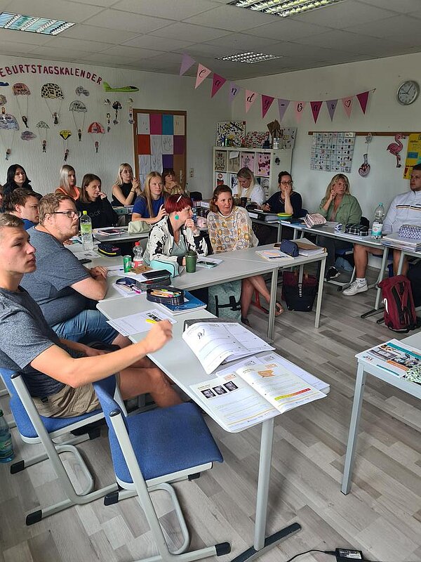 Auszeichnung „Clever in Sonne und Schatten“ 2024 Die Schüler*innen im gut gefüllten Klassenzimmer lauschen gespannt den Referent*innen, die nicht im Bild sind. Der Raum ist bunt geschmückt mit Girlanden und einem Geburtstagskalender an der hinteren Wand. Die Wanduhr zeigt 9:05 Uhr an.