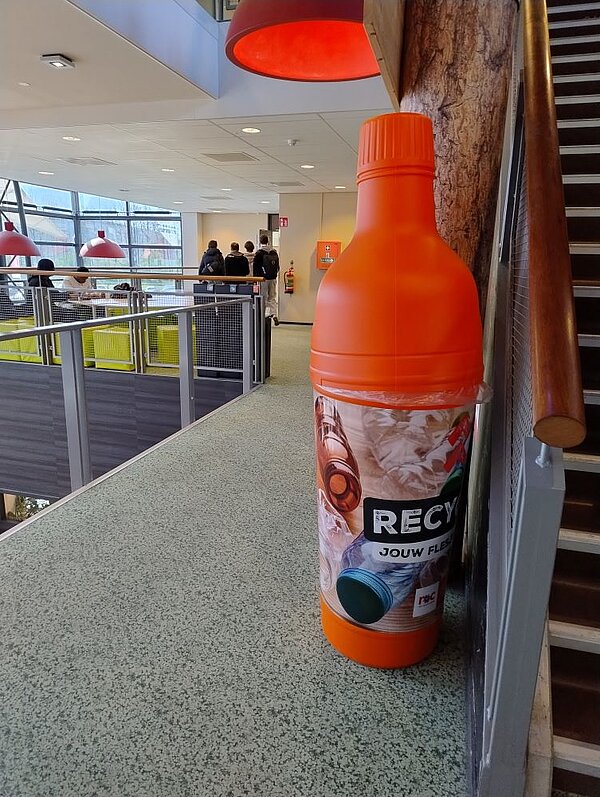Eine große, orangefarbene Flasche als Dekorationselement in einem öffentlichen Gebäude. Die Flasche ist mit einer Folie bedruckt, die an ein Recycling-Symbol erinnert.