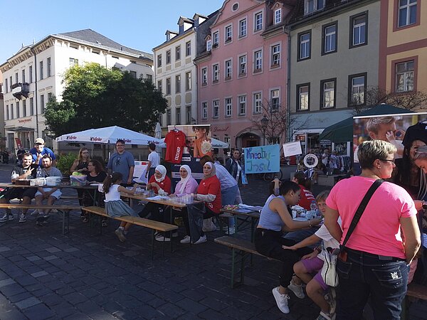 Gruppenfoto am Bastelstand mit vielen Kindern und Erwachsenen an Tischen, dahinter bunte Häuser und Infobanner, auf denen „Erzieher/in“ und „Kinderpfleger/in“ zu lesen ist.