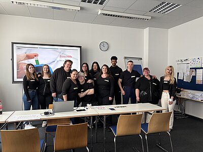 Eine Gruppe von zwölf Auszubildenden in dunkler Kleidung steht vor der digitalen Tafel, auf der eine Präsentation zu Erster Hilfe bei Über- &  Unterzucker läuft.