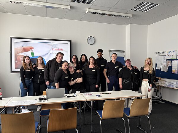Pflegemesse Diabetes an der Euro Akademie Eine Gruppe von zwölf Auszubildenden in dunkler Kleidung steht vor der digitalen Tafel, auf der eine Präsentation zu Erster Hilfe bei Über- & Unterzucker läuft.