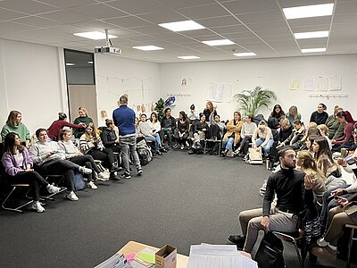 Alle Auszubildenden sind im großen Raum versammelt, sitzen mit ihren Stühlen an den Wänden, sodass in der Mitte Raum entsteht.