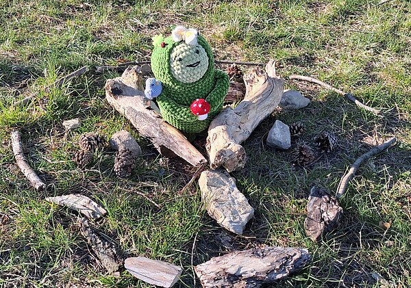 Grüne gehäkelte Figur mit Blume und Pilz, umgeben von Holzstücken, Steinen und Tannenzapfen auf einer Wiese.