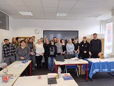 Mehrere Auszubildende stehen in einem Klassenraum in der Euro Akademie Berlin nebeneinander vor einer großen Tafel. Im Vordergrund sind Tische mit verschiedenen Gegenständen wie Flaschen, Papierstapeln und Ordnern zu sehen. Auf einem Tisch liegt ein blaues Tuch mit weißem Aufdruck. Die Wände sind weiß, an der Decke befinden sich mehrere Leuchten, und eine Uhr hängt an der Wand.