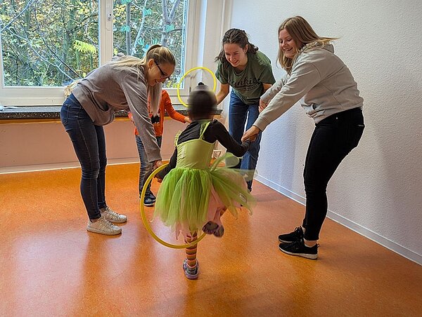 Bewegungsfreude in der Kita Drei weibliche Auszubildende bilden mit zwei Kinder aus der Kita einen Kreis und üben mit einem Hula Hoop Reifen.