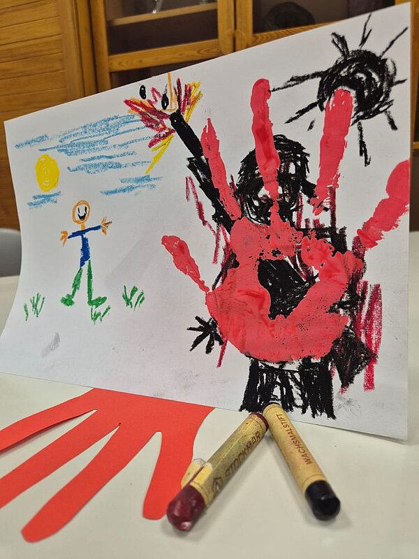 Eine Kinderzeichnung mit einem großen roten Handabdruck in der Mitte auf einer schwarzen Skizze einer Figur, die eine Waffe hält und einer schwarzen Sonne. Links daneben ist eine kleine Figur mit einem lächelnden Gesicht und einer Sonne im Hintergrund. Auf dem Tisch liegen zwei Wachsmalstifte und ein roter Papp-Handabdruck.