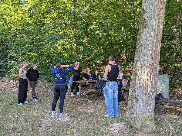 Eine Gruppe von Menschen in einem Wald. Einige stehen um einen Picknicktisch herum, andere posieren für ein Foto. Die Szene ist von dichtem Grün und Bäumen umgeben.
