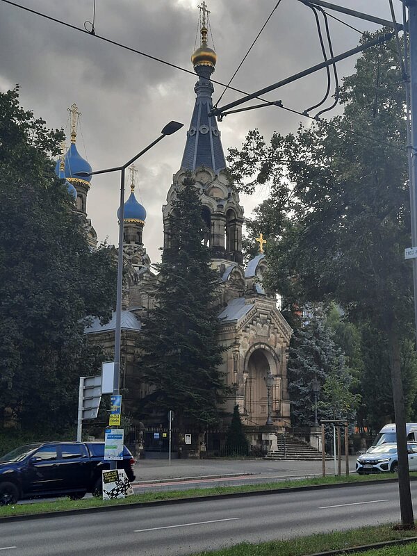 Ethikunterricht mal anders Straßenszene mit Blick auf eine orthodoxe Kirche mit blauen und goldenen Zwiebeltürmen, umgeben von Bäumen. Im Vordergrund sind Autos auf einer mehrspurigen Straße sowie Straßenlaternen und Oberleitungen zu sehen.