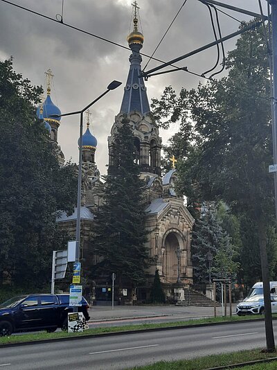 Straßenszene mit Blick auf eine orthodoxe Kirche mit blauen und goldenen Zwiebeltürmen, umgeben von Bäumen. Im Vordergrund sind Autos auf einer mehrspurigen Straße sowie Straßenlaternen und Oberleitungen zu sehen.