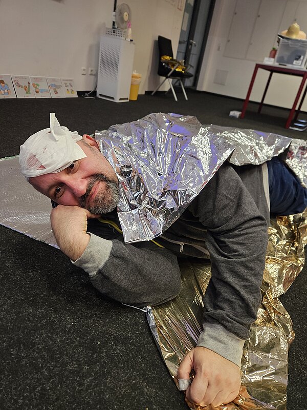 Teilnehmer mit Kopfverband und Rettungsdecke liegt in stabiler Seitenlage und blickt zur Kamera; Schulungsposter und Materialien im Raum.