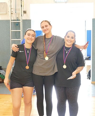 Drei Frauen stehen nebeneinander in einem Innenraum und tragen Medaillen um den Hals. Sie lächeln und posieren für ein Foto. Die Frau in der Mitte trägt ein graues T-Shirt, während die anderen beiden schwarze Sportkleidung tragen.