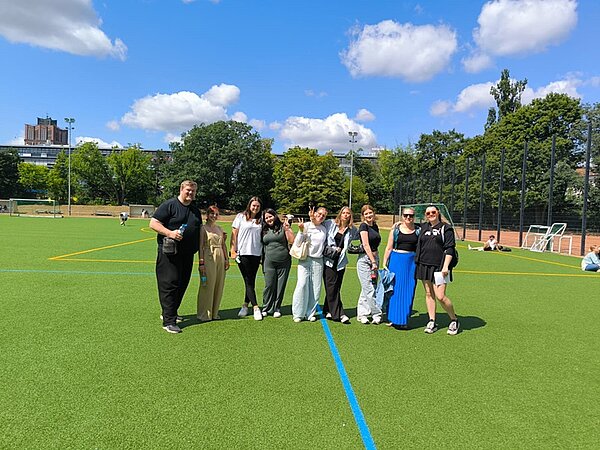 Schnitzeljagd, Tombola und Volleyball-Turnier Auszubildende der Euro Akademie Berlin stehen auf dem großen Rasen-Sportplatz, am strahlend blauen Himmel herrliche Cumuluswolken, und posieren für ein Gruppenfoto.