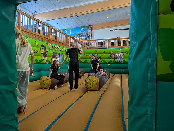 Eine große Indoor-Hüpfburg mit gelbem Boden und grünen Wänden, auf denen Lianen und Blätter sowie ein Löwe, ein Affe und ein Elefant abgebildet sind. Eine Gruppe junger Menschen sitzt und steht im Inneren der Hüpfburg um zwei Bänke, die wie Baumstämme geformt sind.