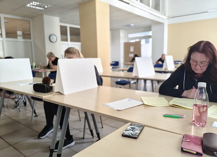 Die Prüflinge sitzen in einem großen Raum an Tischen vor ihren Abschlussprüfungen, es sind Sichttrenner zwischen ihnen aufgestellt, auf den Tischen liegen Stifte und Wasser, die Prüflinge schreiben konzentriert ihre Prüfung. 