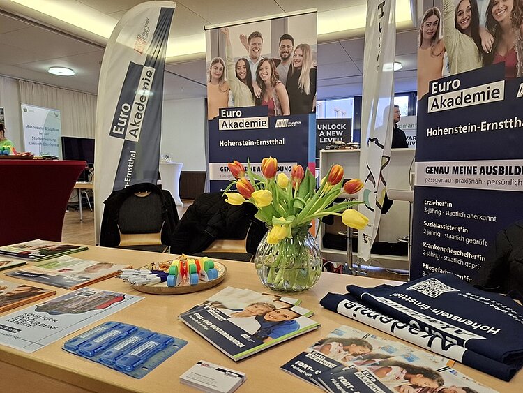 Messestand der Euro Akademie Hohenstein-Ernstthal mit Flyern, Broschüren, Werbegeschenken, einem Tulpenstrauß und zwei großen Roll-ups mit Logos und Informationen zur Ausbildungsmesse in der Stadthalle Limbach-Oberfrohna.