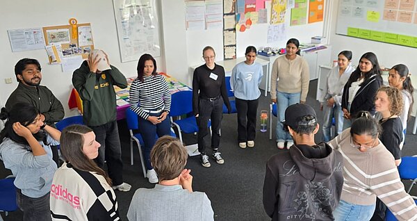 Teilnehmende stehen in einem Klassenraum im Kreis und tauschen sich gemeinsam zu einem Workshop-Thema aus.