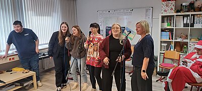 Sechs junge Menschen und eine Lehrkraft singen gemeinsam mit Mikrofonen Karaoke in einem Klassenraum, im Hintergrund sitzt eine als Weihnachtsmann verkleidete Person auf einem Stuhl.
