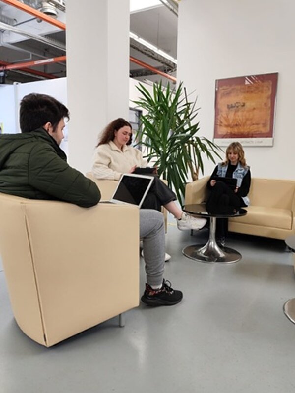 Drei Auszubildende sitzen im hellen, großen Foyer der Euro Akademie Nürnberg auf hellen Sesseln und Sofas, mittig ein Tisch, und haben Notebooks auf dem Schoß.